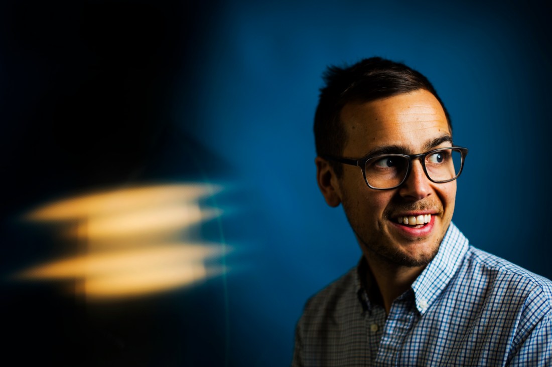 A man wearing glasses stands against a blue background and looks over one shoulder, smiling.