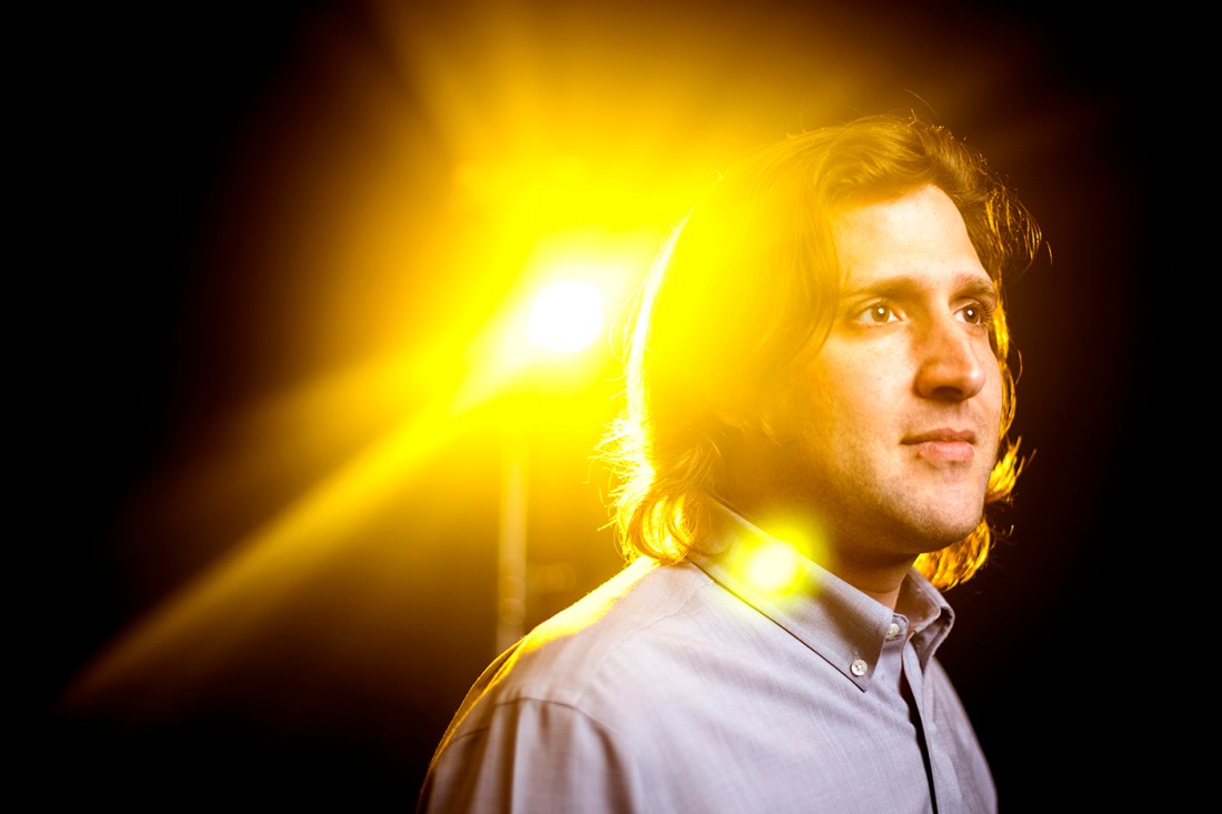 John Basl, a professor of philosophy, has collar-length hair and a collared shirt. A bright yellow light flashes in the background.