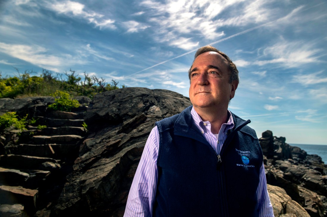 Portrait of H. William Detrich wearing a striped shirt under a vest with a natural landscape and blue sky in the background. 