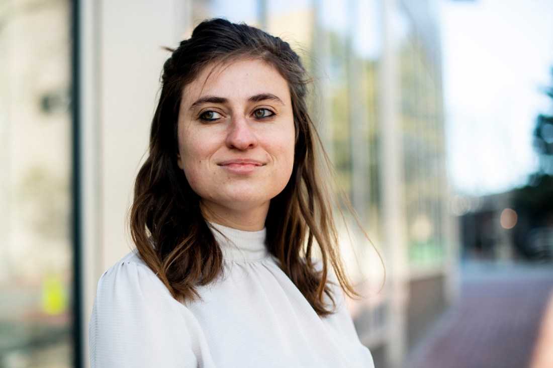 Cassie McMillan, a young woman with brown hair and an assistant professor of sociology, looks off into the distance in front of a storefront.