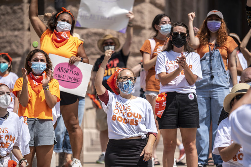 pro-choice protesters in austin yelling and raising their firsts while wearing orange and white shirts that say 