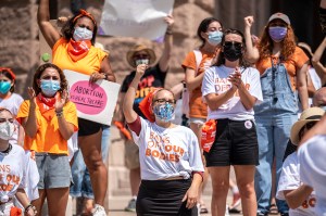 pro-choice protesters in austin yelling and raising their firsts while wearing orange and white shirts that say "bans off our bodies"