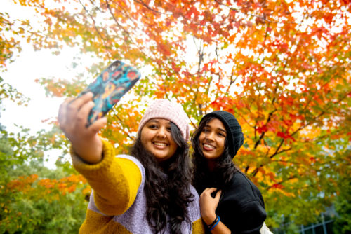 Kimaya Khilare and Devashree Kulkarni, both graduate students studying information systems, take a selfie under a tree full of foliage on Northeastern’s Boston campus on Oct. 26, 2021. Photo by Matthew Modoono/Northeastern University