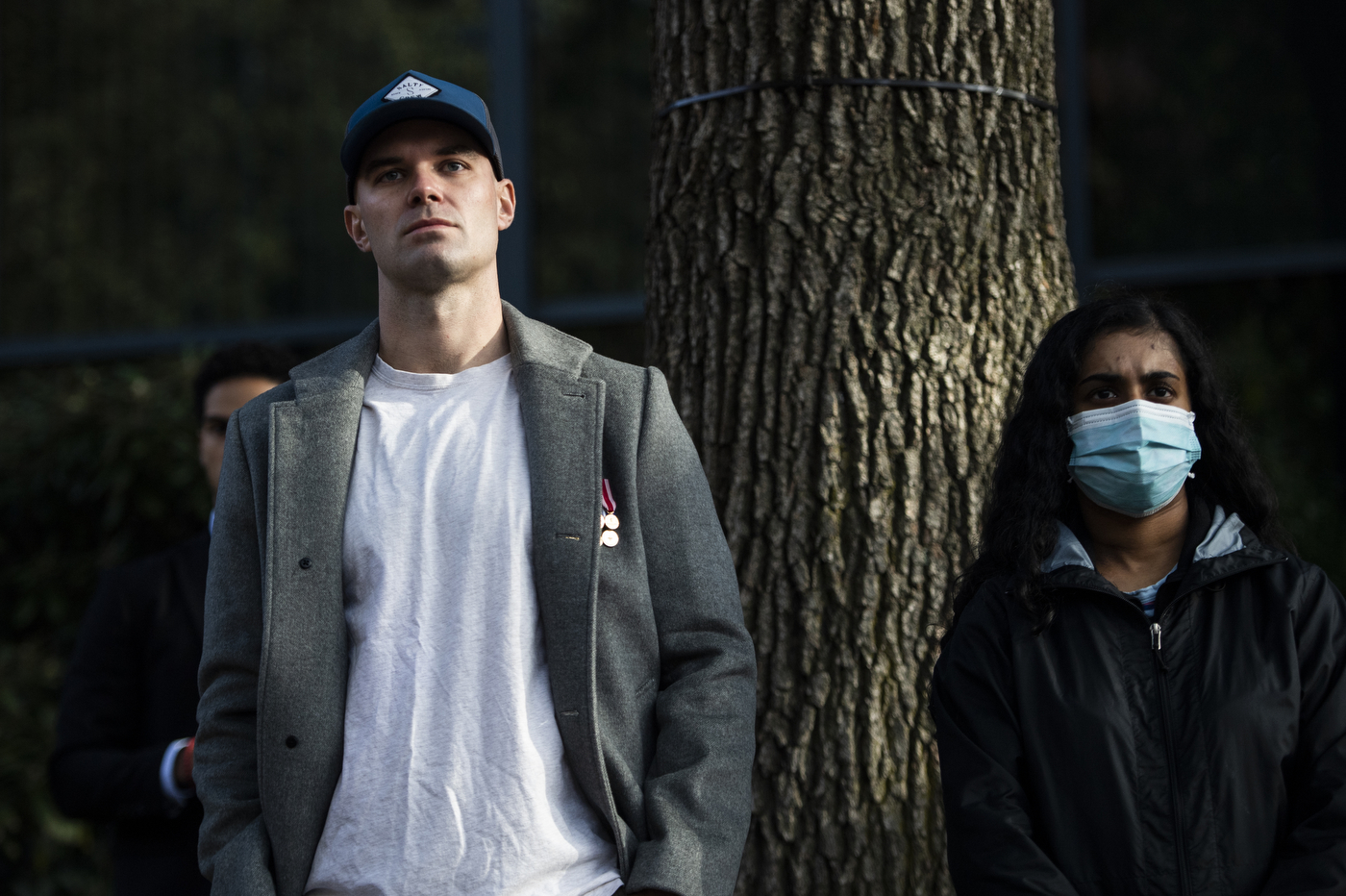 man with grey jacket and blue baseball cap stands next to a masked woman