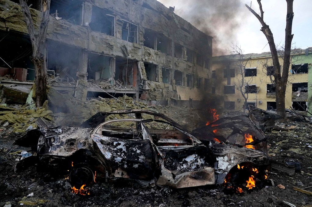 A mangled, burning car in front of war-damaged buildings