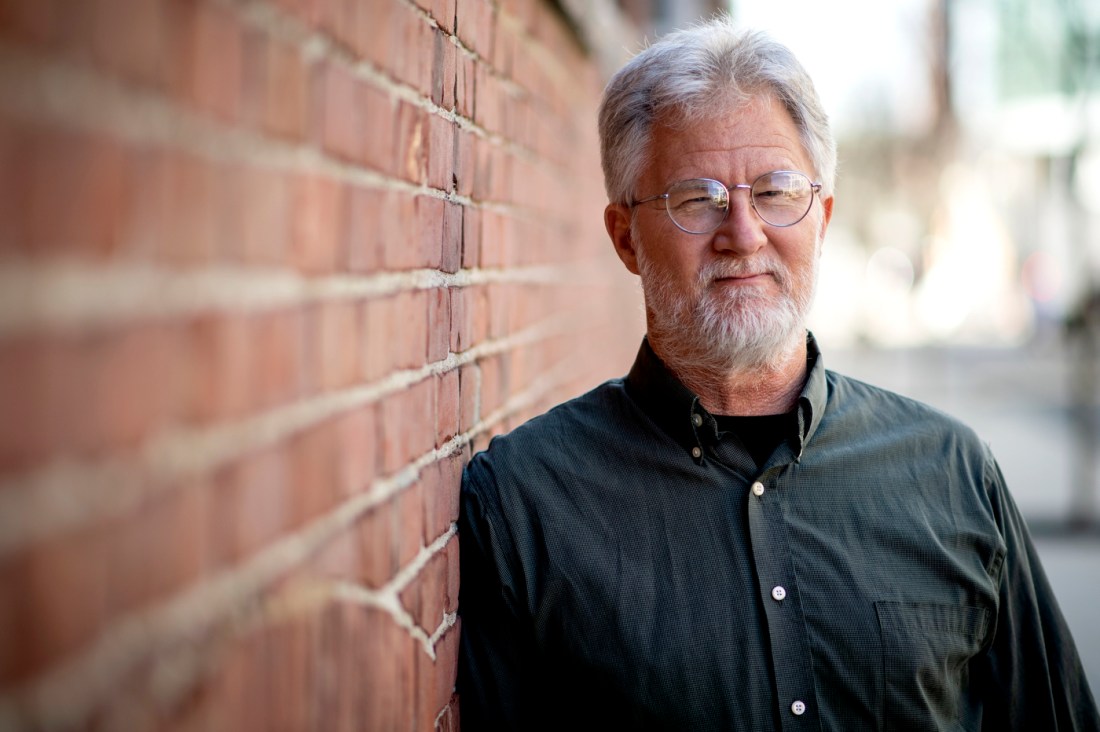 A man with gray hair and glasses stands against a red-brick wall.