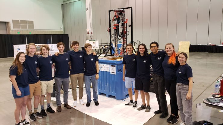 Eleven students in matching t-shirts standing in a large room, smiling proudly in front of a large machine
