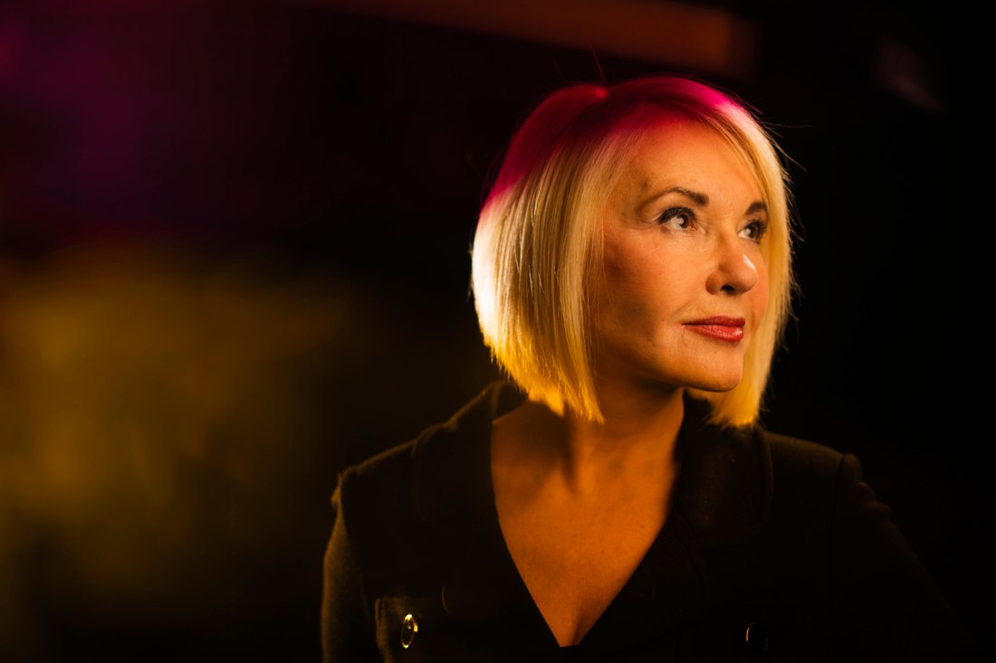 A woman with blonde hair dressed in black looks to her left. She has a neutral expression.