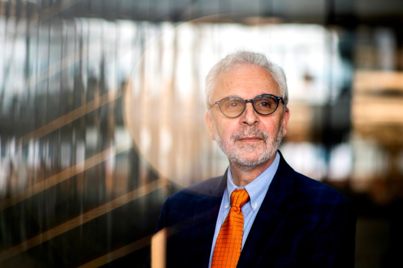 Nikos Passas, wearing glasses, a dark suit, and an orange tie, in a professional portrait with a softly blurred architectural background.