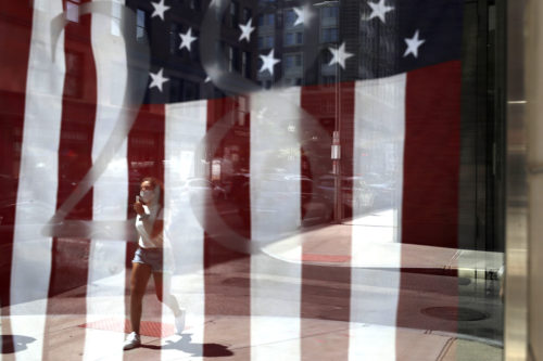 A person wearing a mask to help prevent the spread of the coronavirus walks past a U.S. flag displayed in a window in Boston on July 7, 2020. AP Photo/Steven Senne