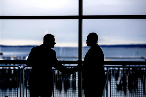 A silhouette of two men shacking hands against the background of windows facing the ocean.
