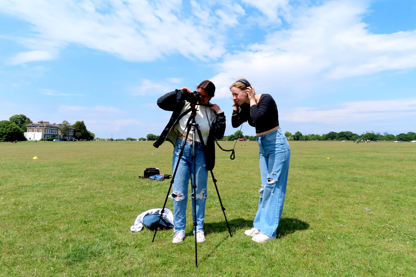 Students filming on an open green field; one is looking through the camera and the other is listening through headphones