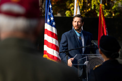 man wearing suit and tie speaking at a podium with a united states and military flags behind him