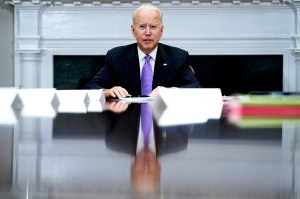white haired man wearing purple tie sitting at a large desk