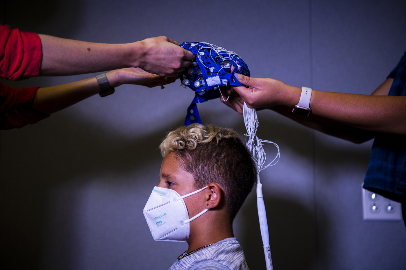 A study participant is pictured as two researchers lower a cap with sensors and wires onto their head