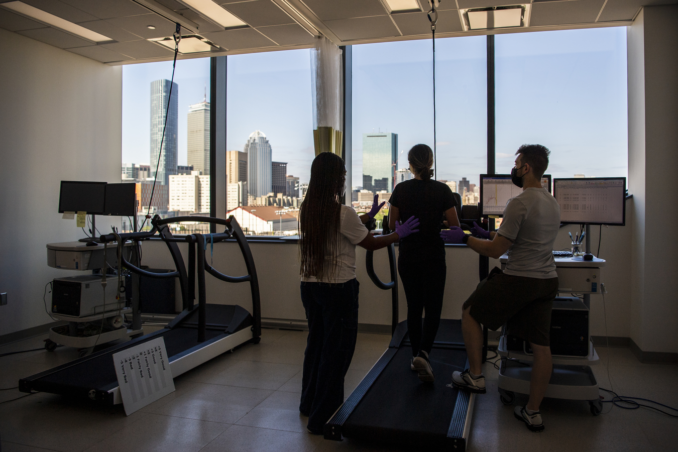 A study participant runs on a treadmill while two researchers look on. The treadmill faces a window with a view of the Boston skyline. 