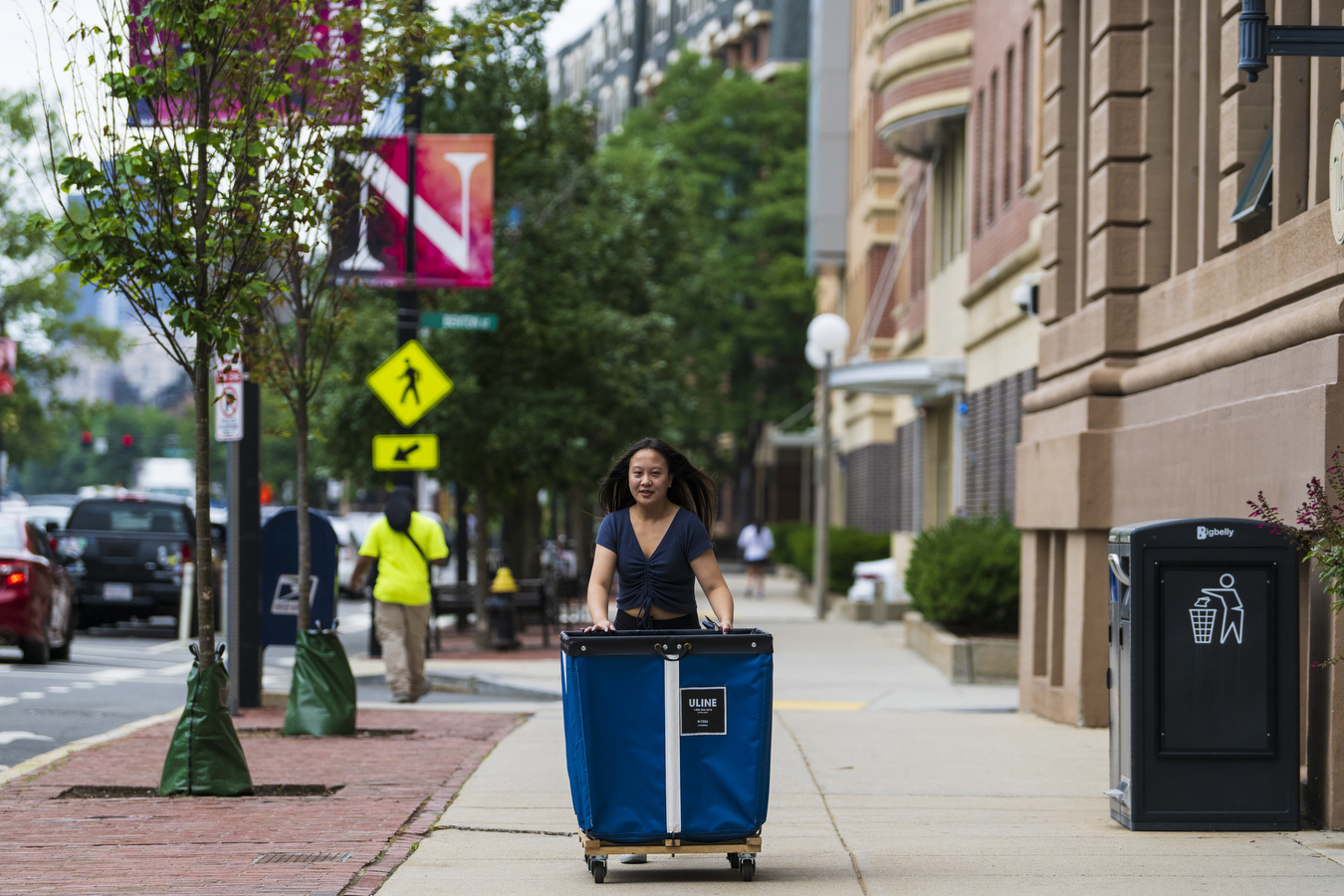 A person walks down the sidewalk towards the camera, wheeling an oversized moving bin. 