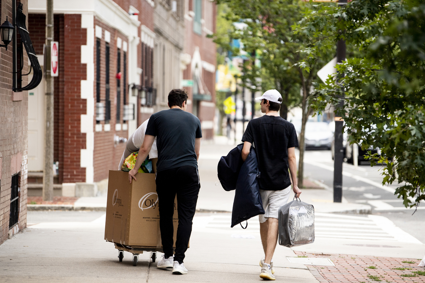 Two students are pictured from the back, walking down the sidewalk. Once student (left) has a wheeled-cart full of dorm-room items. One student (right) is carrying a garment bag in one arm and a small bag in the other. 