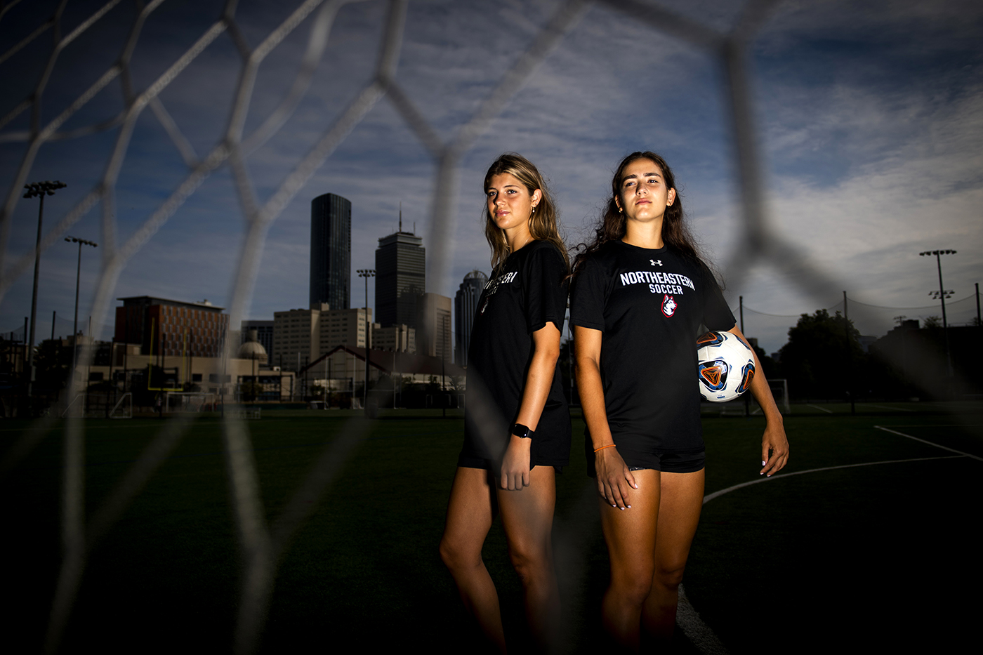 Two soccer players pose against a Boston skyline backdrop.