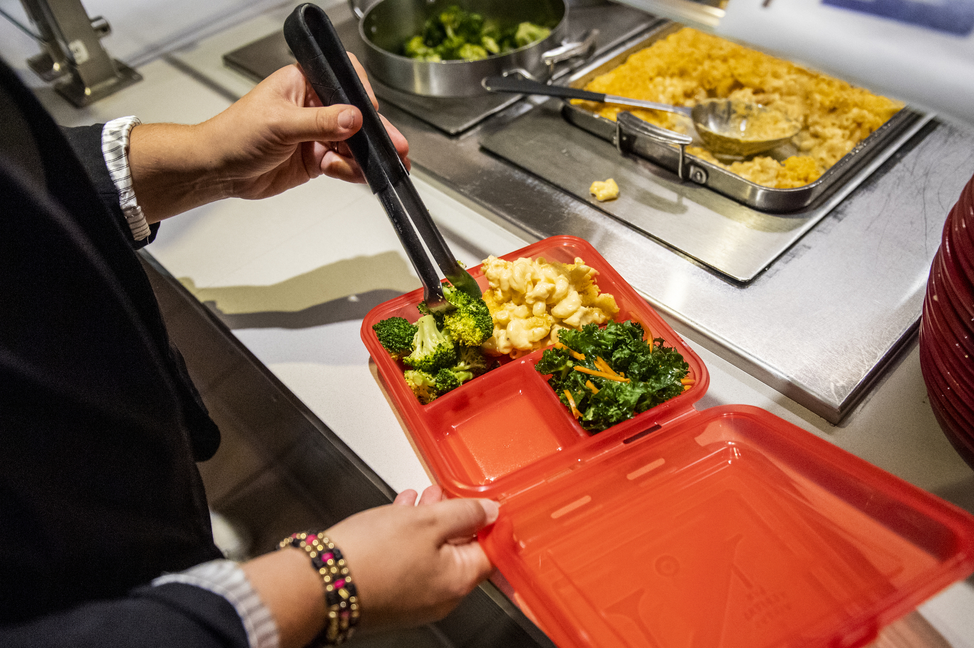 A person is using food prongs to place broccoli inside a red food container.