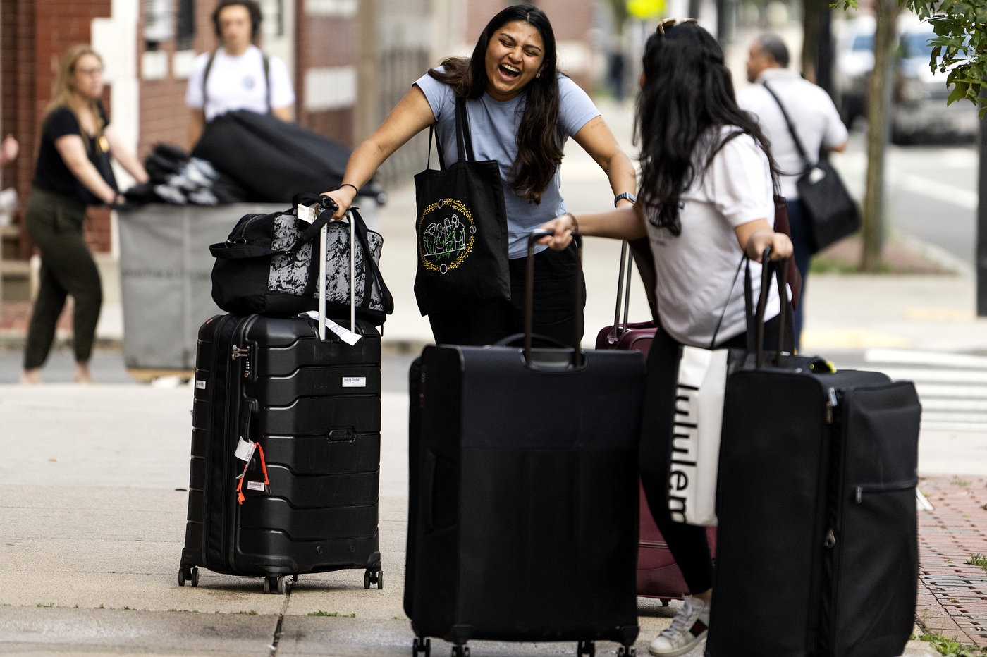 Two students stand on the sidewalk with their luggage. One person is facing forward laughing and the other is turned talking to the other student