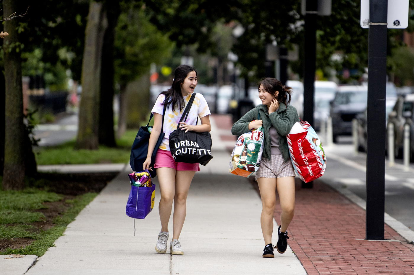 Two students look at each other, laughing, as they walk down the sidewalk, each carrying multiple tote bags in each arm. 