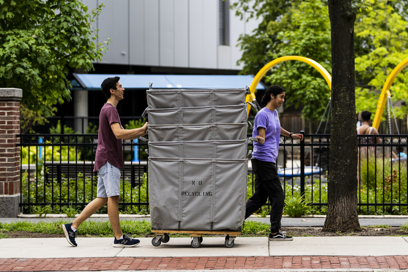 Two people walk down the sidewalk, each helping to guide a stack of oversized moving bins. 