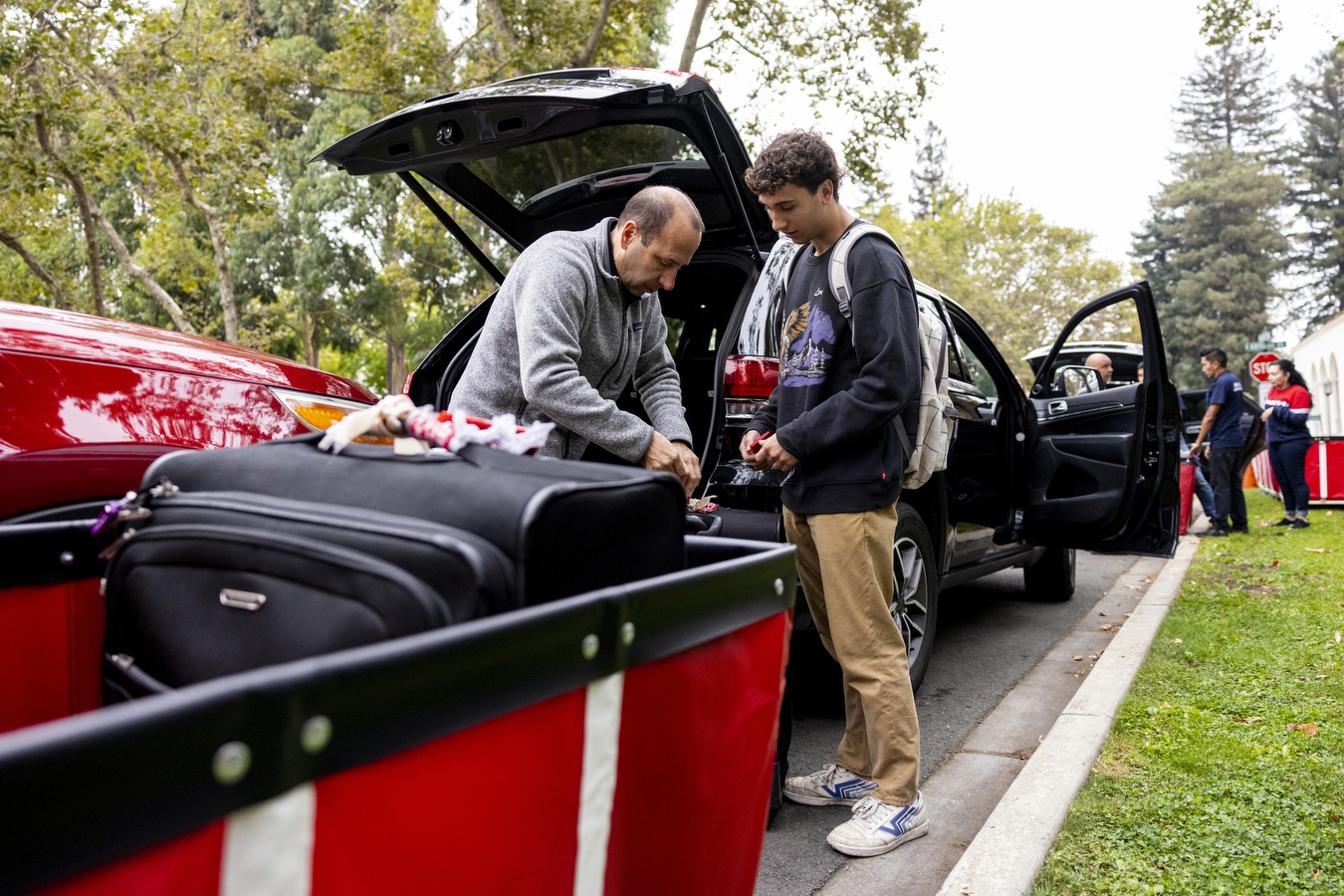 A father helps his sone unload a suitcase and belongings form the back of a black SUV