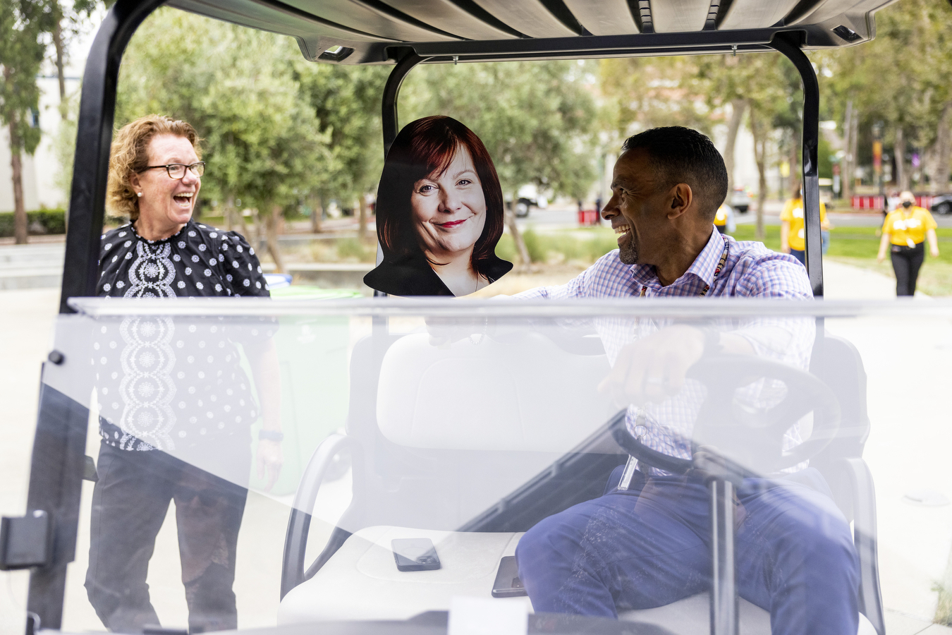A staff member drives a golf cart, smiling at another staff member who looks on, walking, smiling back. Between them is a cardboard cutout of another staff member. 