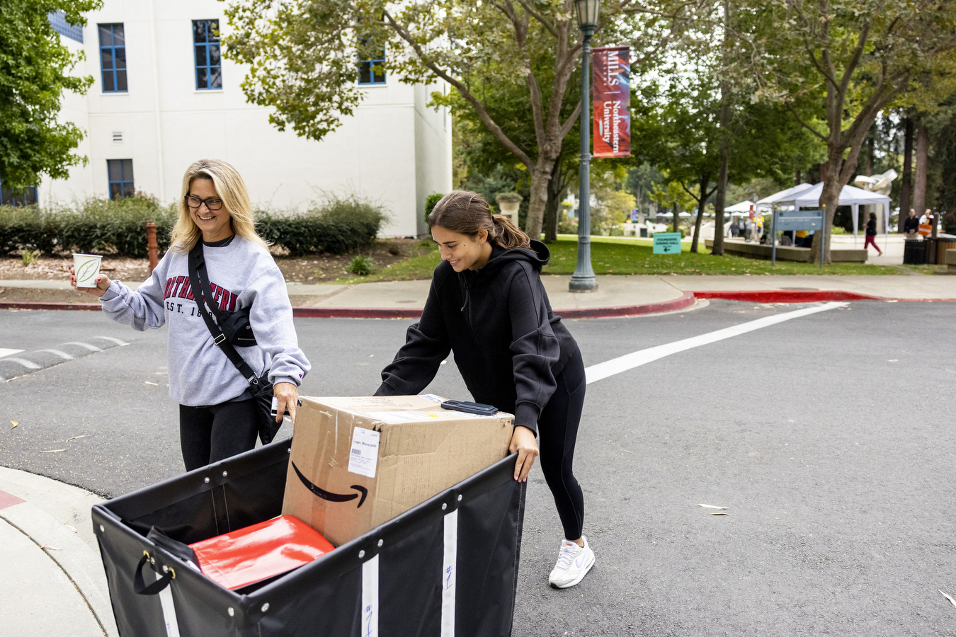 A student is assisted by her mother as she wheels a moving bin full of personal items across a street
