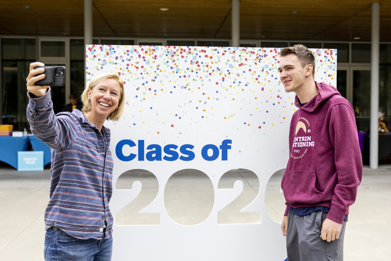 A mother (left) and her son(right) pose for a selfie in front of a white sign with multi-colored confetti with the words "Class of" seen