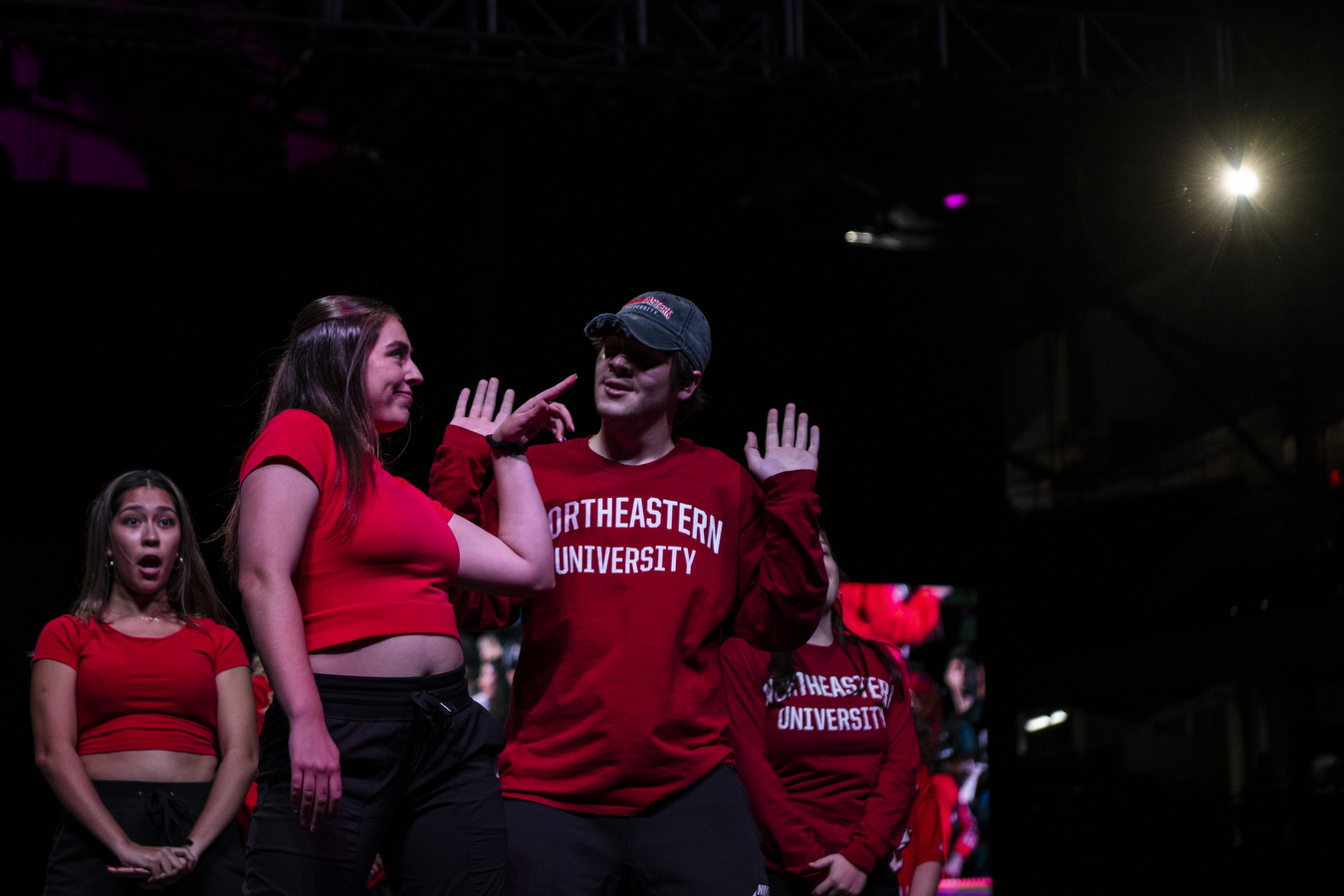 A group of people wear red shirts and black pants while dancing together in a dimly lit arena. 