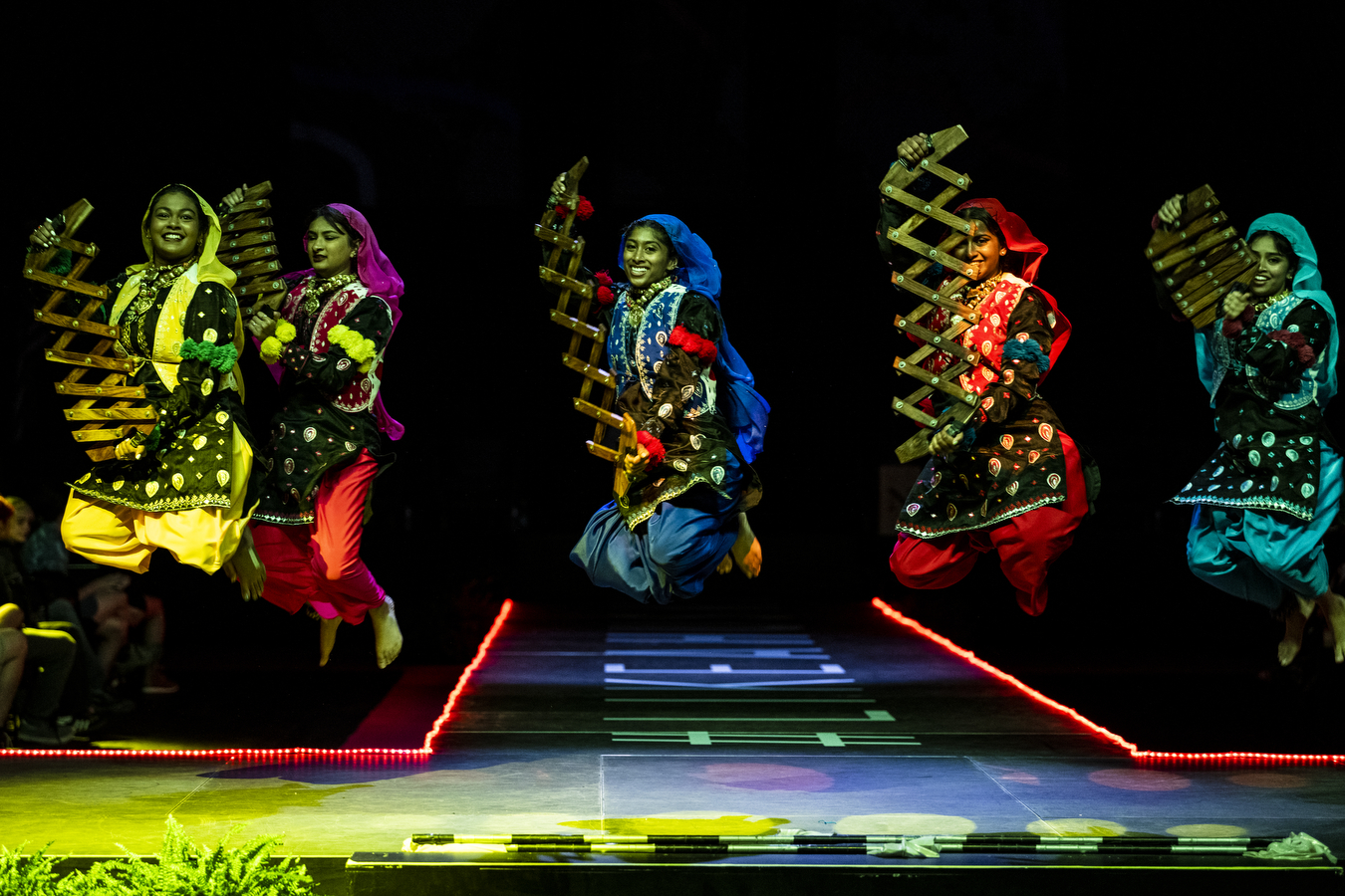 Five people wearing multi-colored outfits are dancing together on a stage while holding large pieces of wooden instruments. 