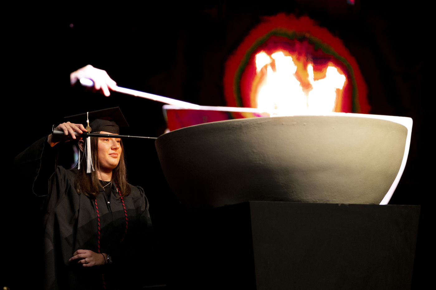 A person wearing a graduation cap and gown is lighting a torch at Northeastern's 2022 Convocation.