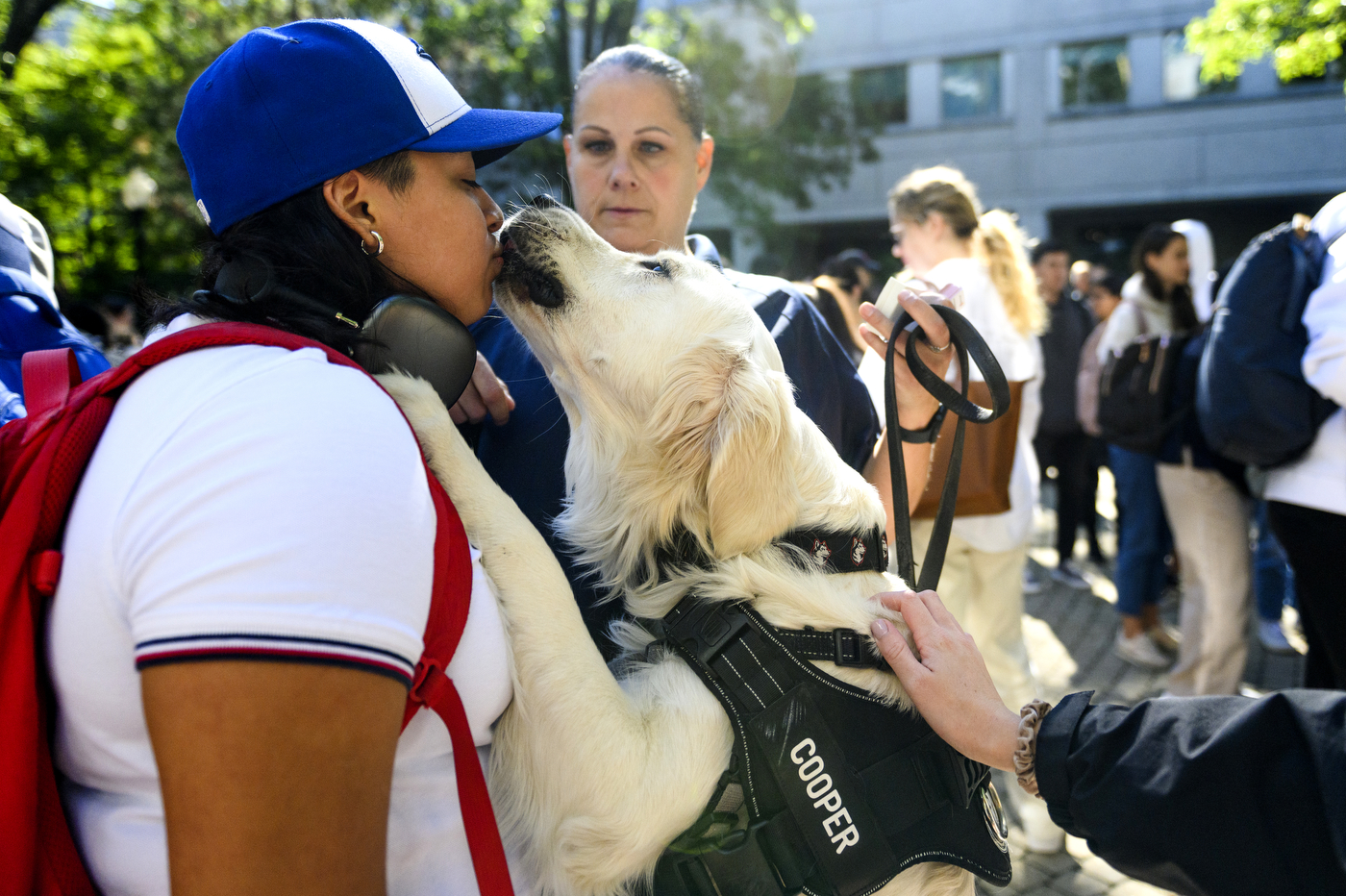 A person wearing a blue hat, white shirt, and red backpack is giving kisses to a white dog. 
