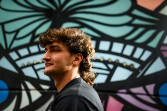 A person with a mullet, wearing a black shirt, stands off to the side near a multi-colored mural.