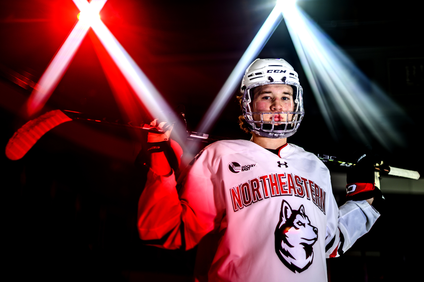 Cam Lundon poses holding a hockey stick over his shoulders wearing a Northeastern hockey jersey and helmet