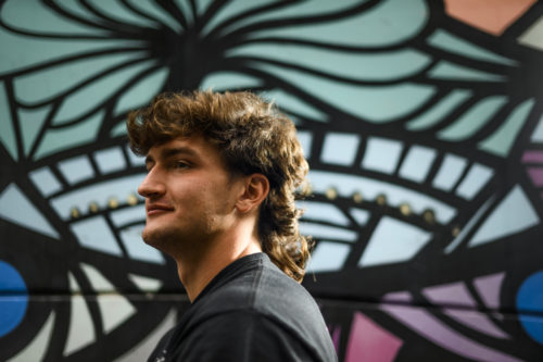 A person with a mullet, wearing a black shirt, stands off to the side near a multi-colored mural.