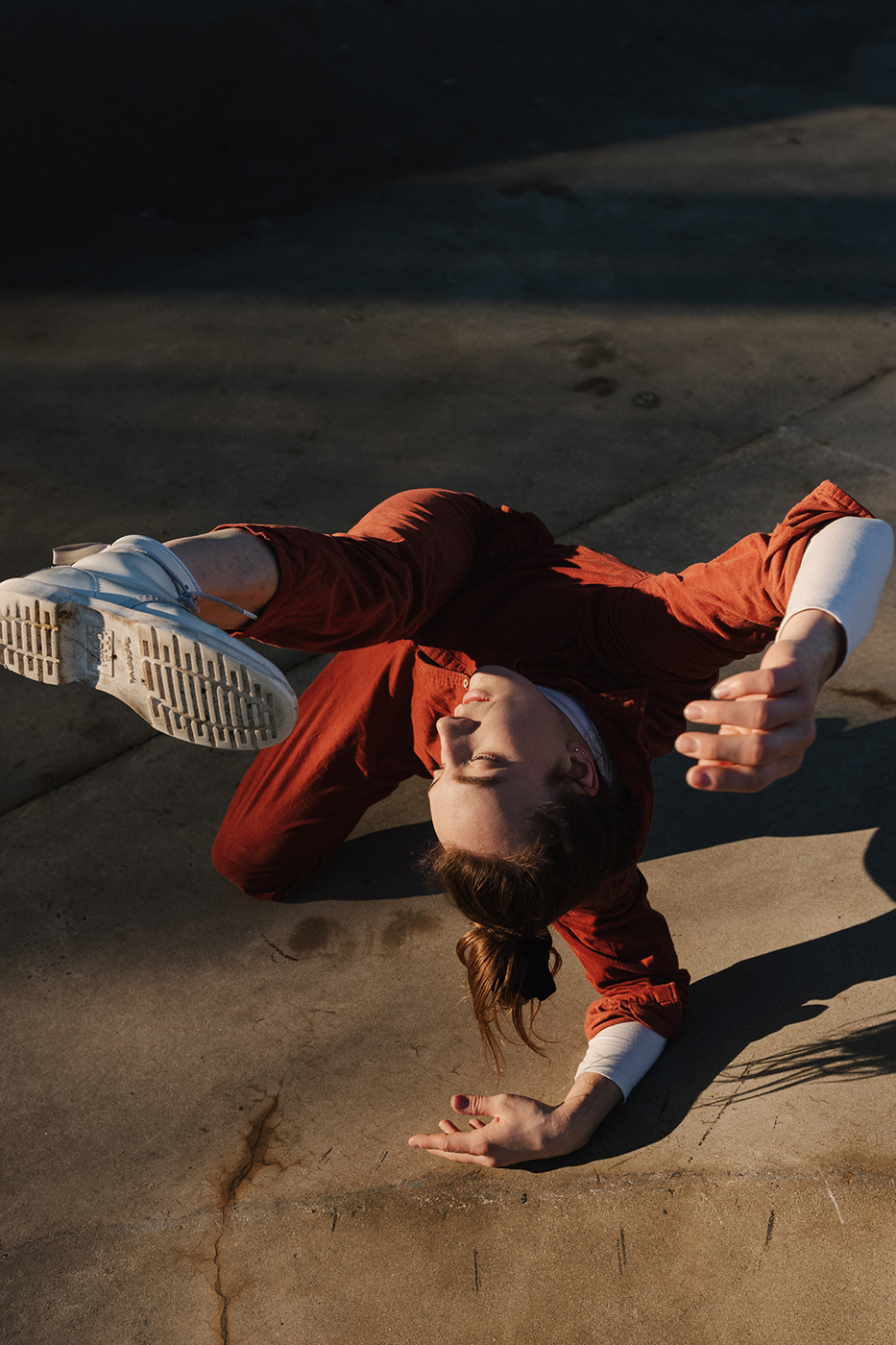 A person wearing a red jumpsuit is dancing on the pavement in the sunlight.