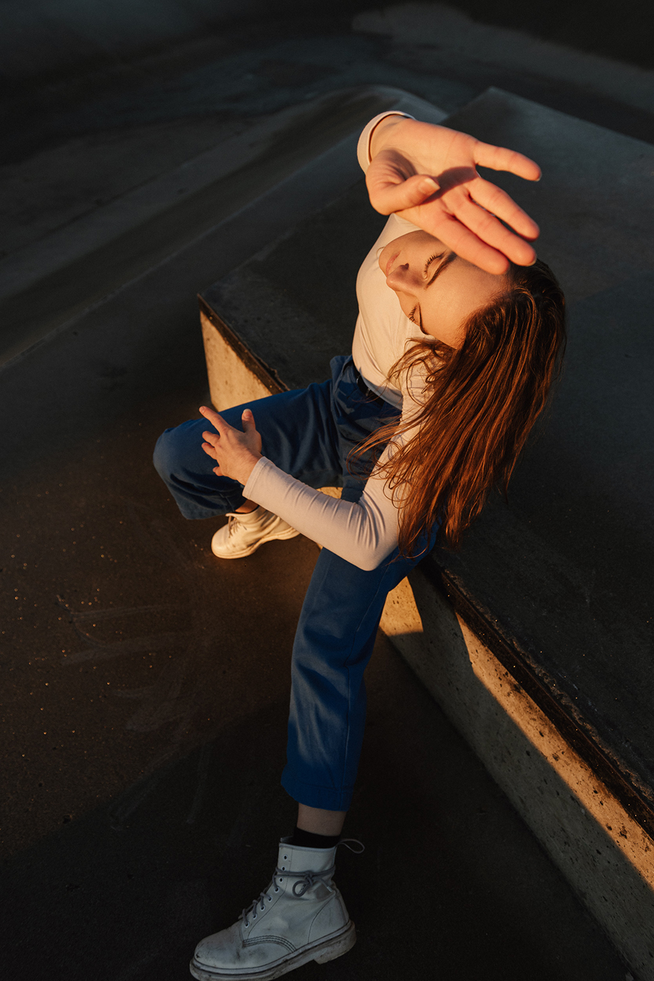 A person wearing a white long-sleeved shirt and jeans is dancing on the pavement in the sunlight.
