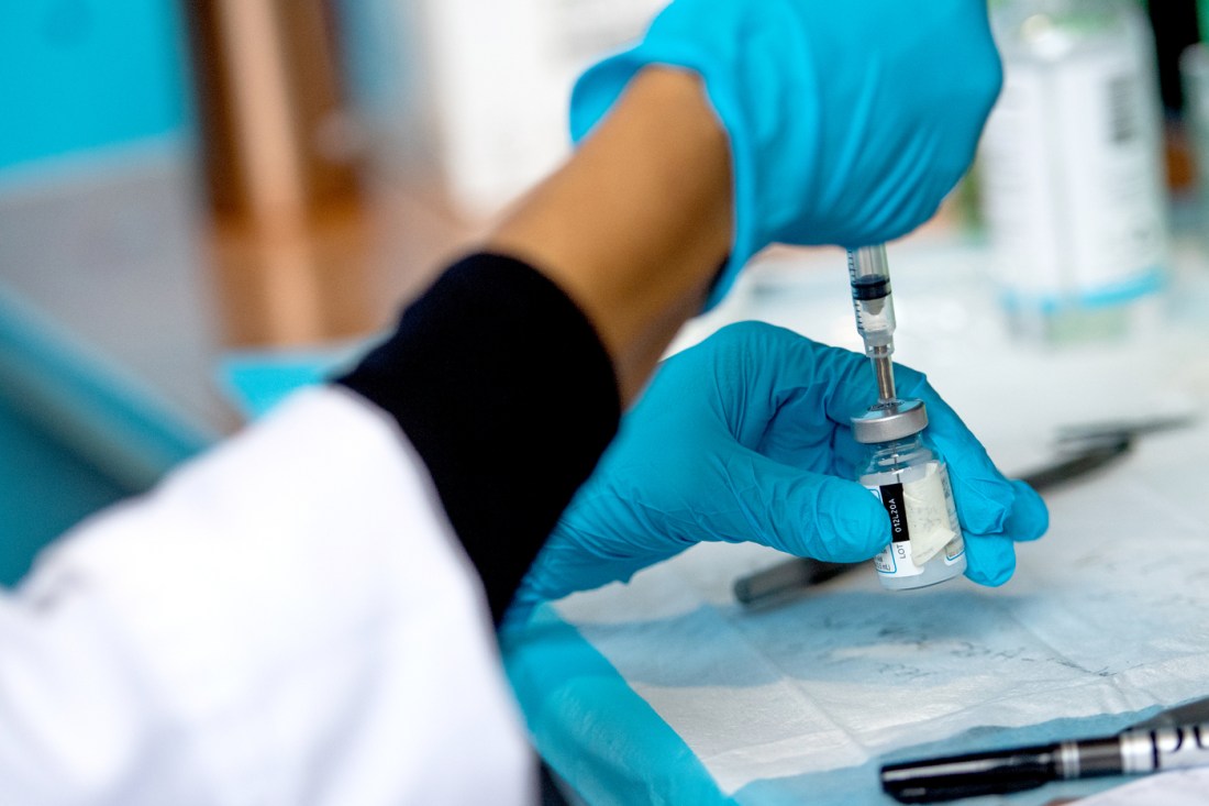 A person wearing medical gloves inserts a needle into a vial of vaccine