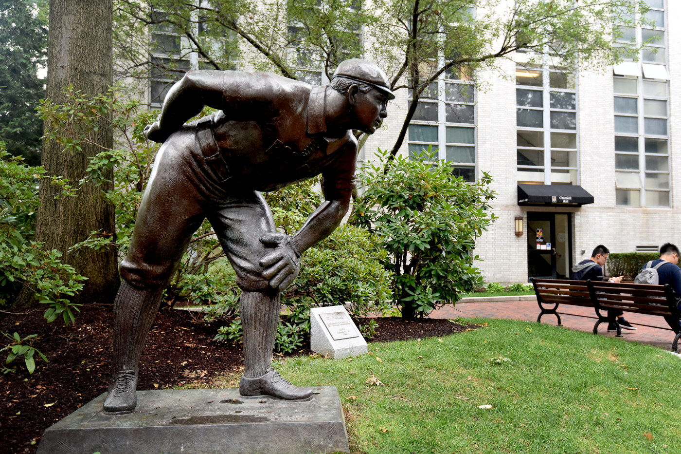 A bronze statue of a baseball player is displayed around bushes and trees on Northeastern's Boston campus. 