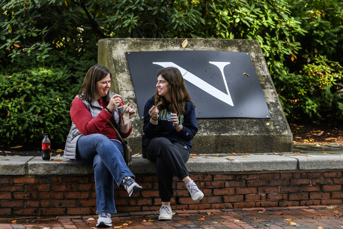 Two people eating ice cream sitting next to a large black sign with the letter 'N' in white text at Northeastern's Family and Friends Weekend.