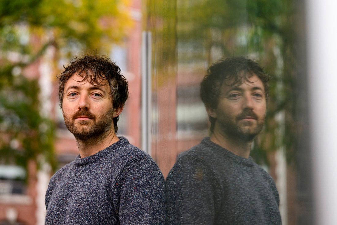 Chad Lee-Stronach has short hair and a beard. He is leaning up against a glass pane and has a neutral expression.