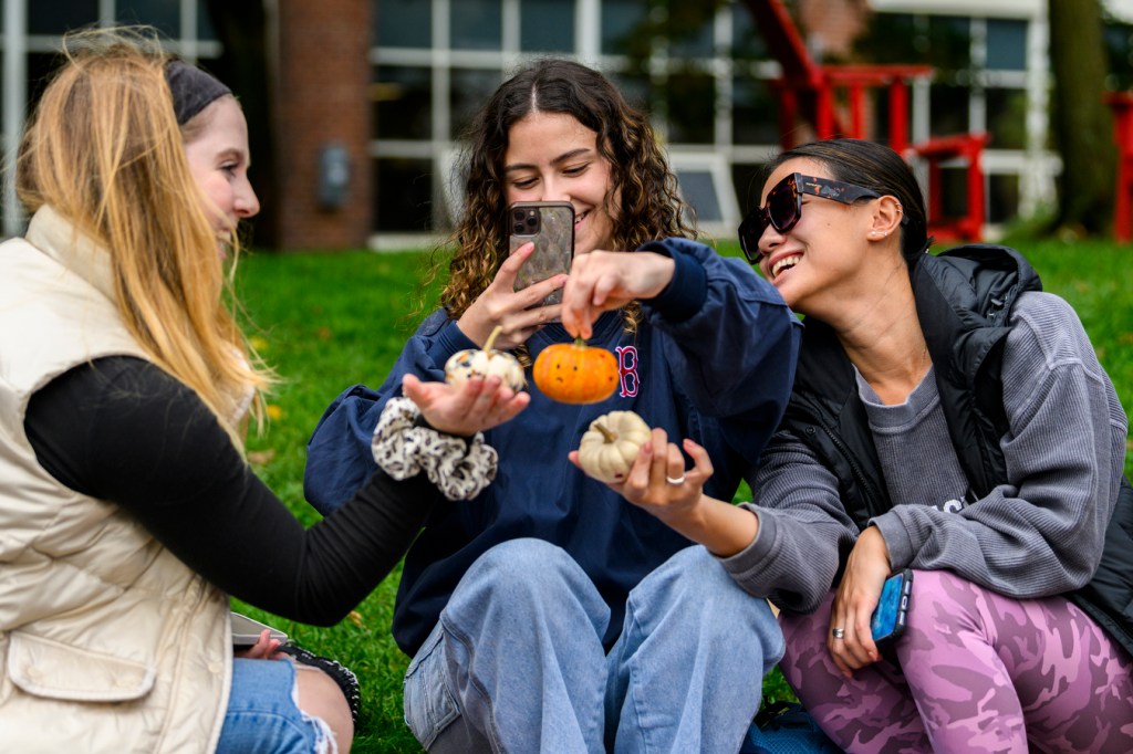 Three people sit in a circle while holding small pumpkins, taking pictures, and laughing.