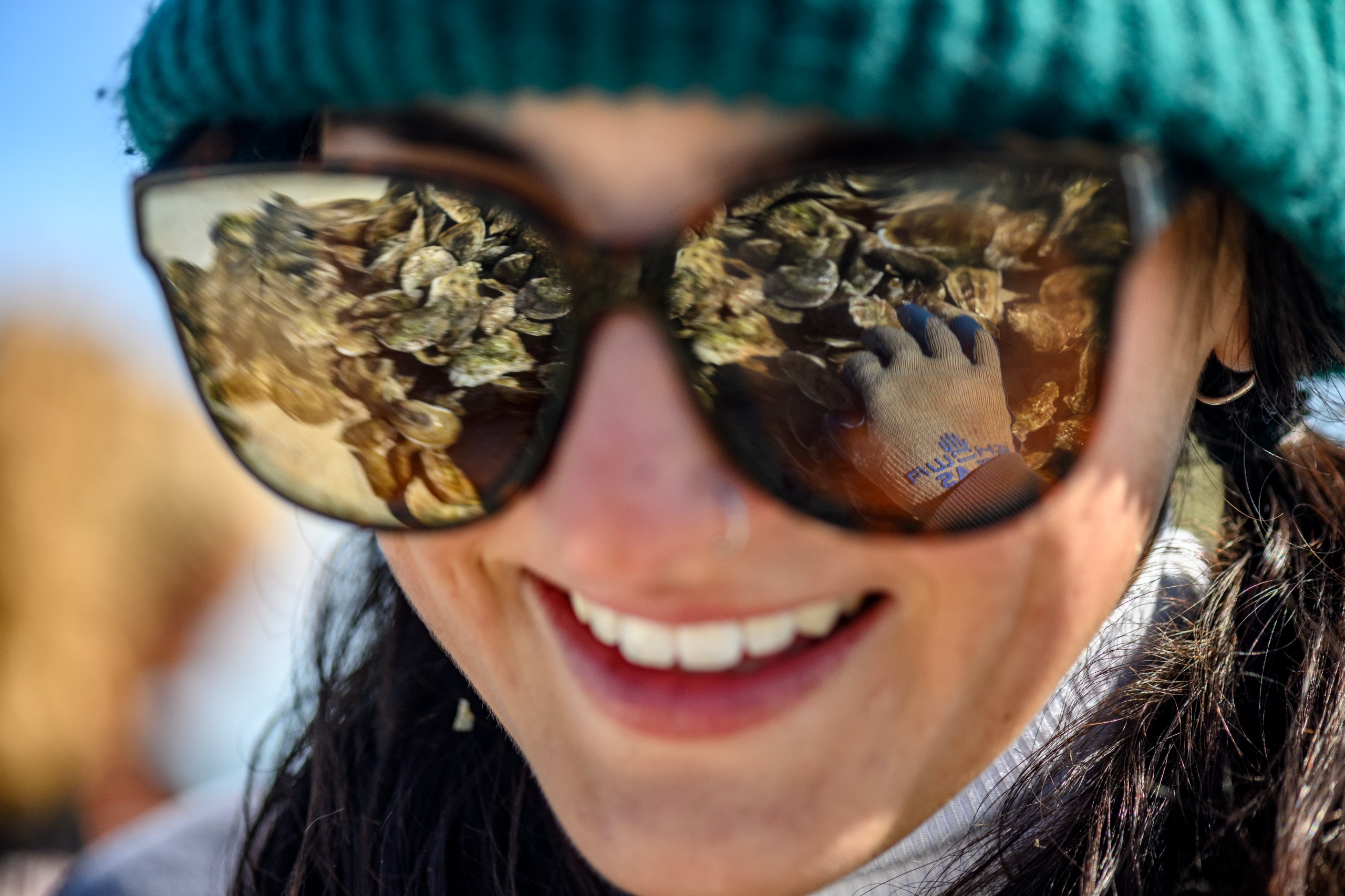 A smiling Nonesuch worker with a reflection of their hand culling oysters in their sunglasses.