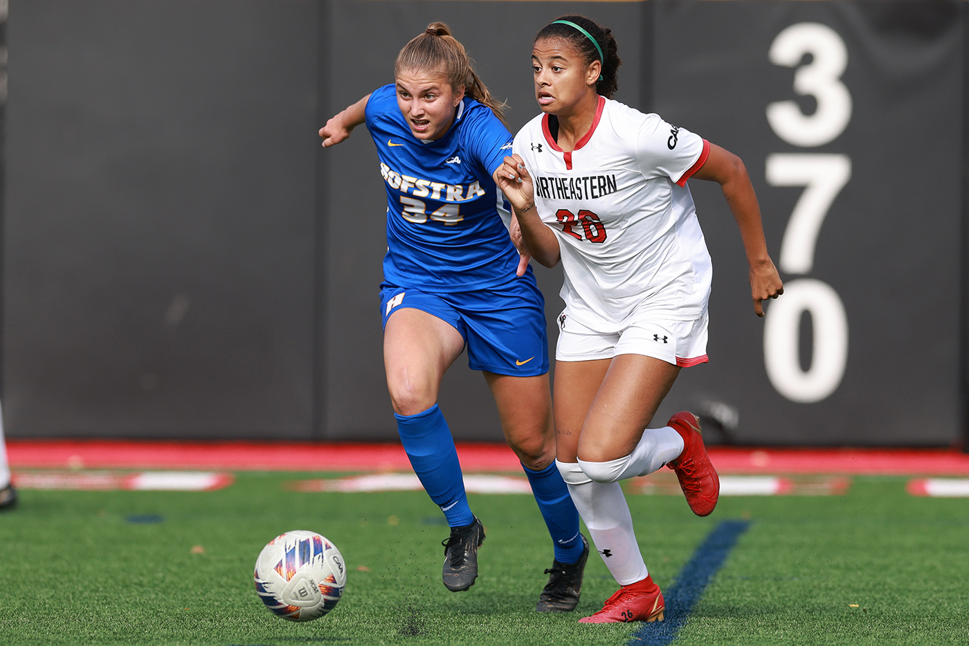 A player on the Northeastern women's soccer team is racing after a soccer ball next to a rival player.