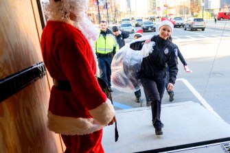 police officer wearing santa hat carrying a bag full of toys past santa claus