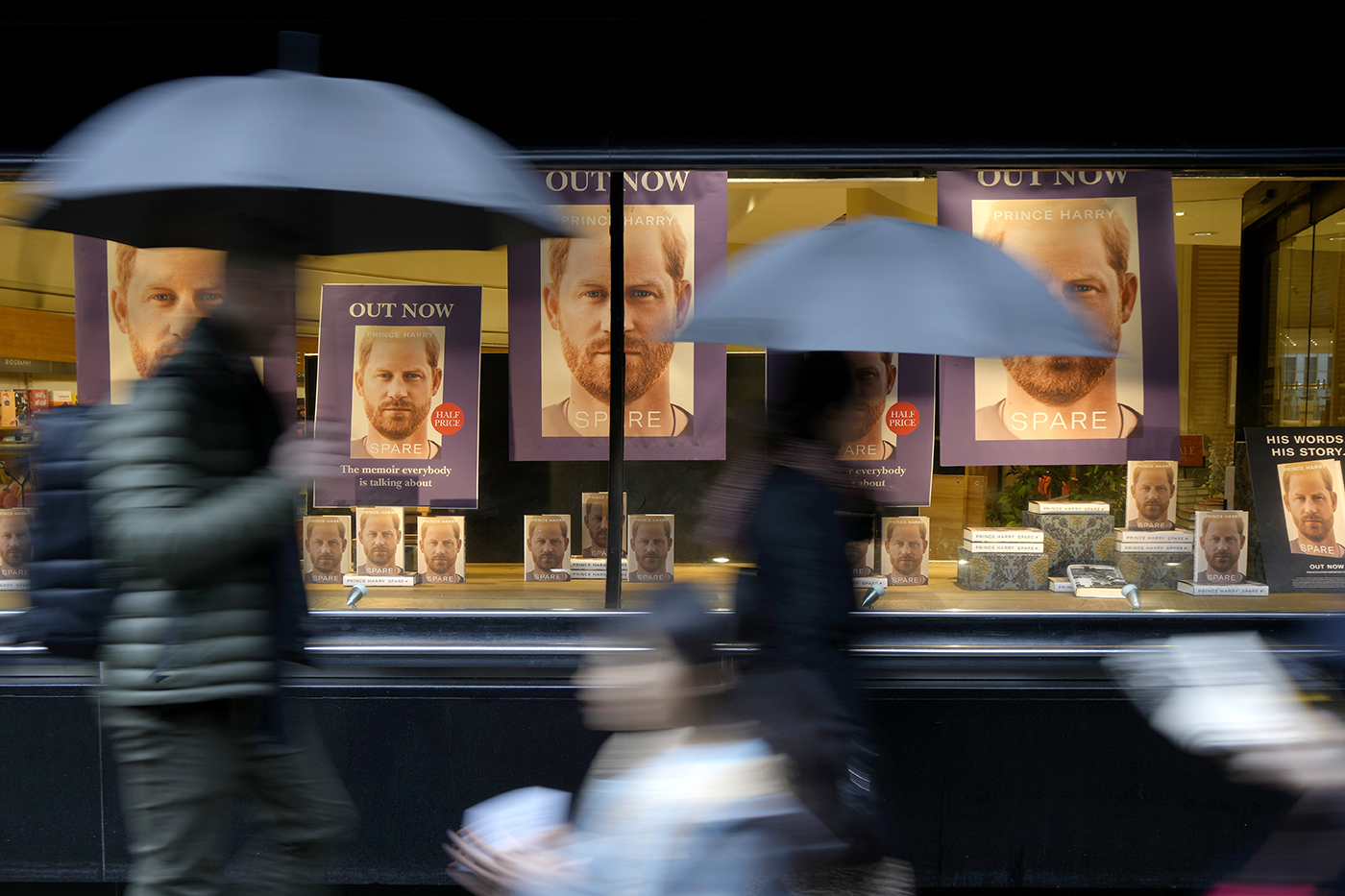 people carrying umbrellas walk past a bookstore window display of prince harry's memoir
