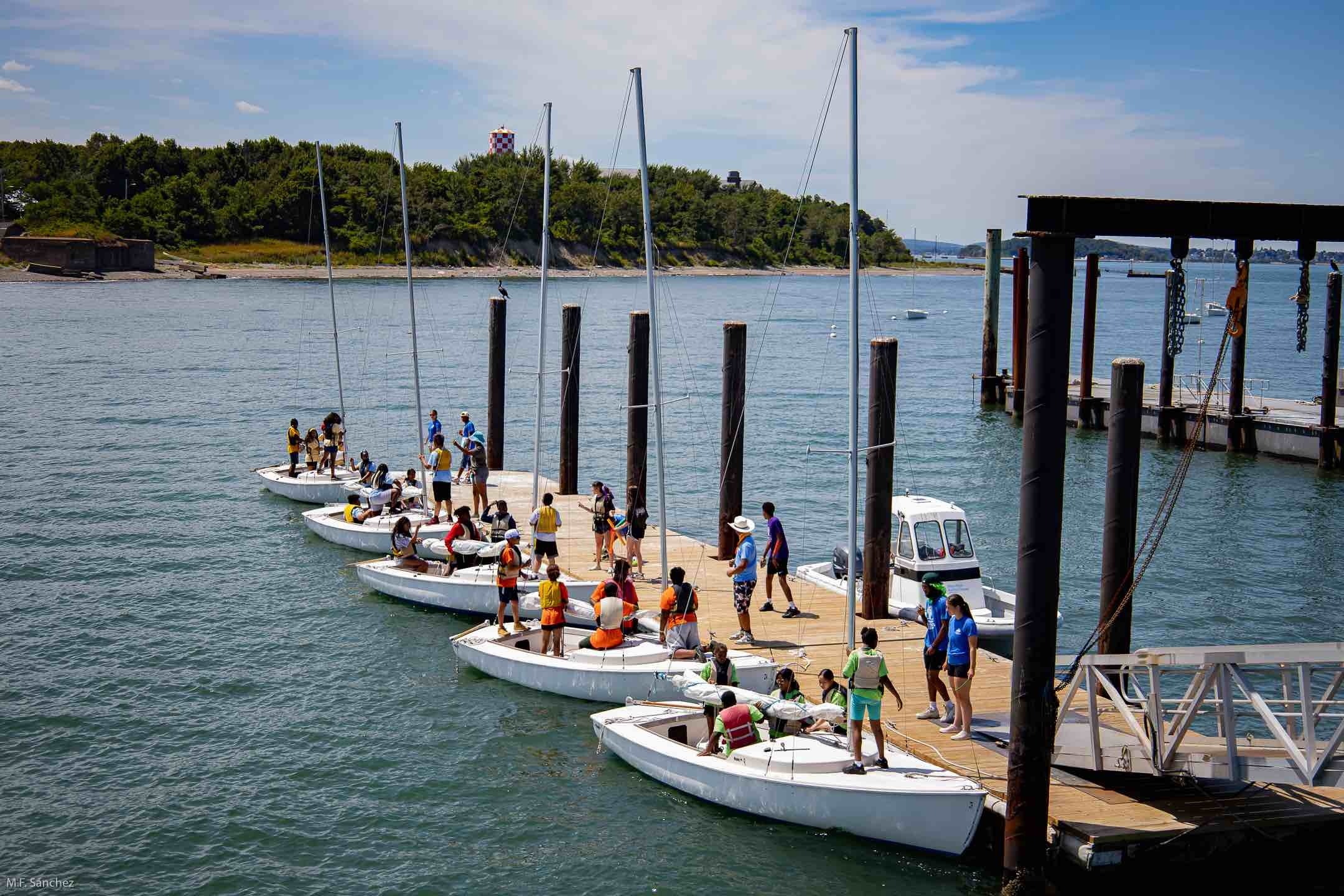 Students at Camp Harbor View getting into sailboats; each boat consists of a team wearing the same color